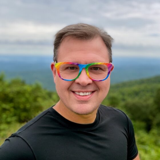 A white man with short brown hair, wearing glasses with a rainbow colored rim. He's wearing a black T-shirt and standing outside with mountains or hills blurred behind him