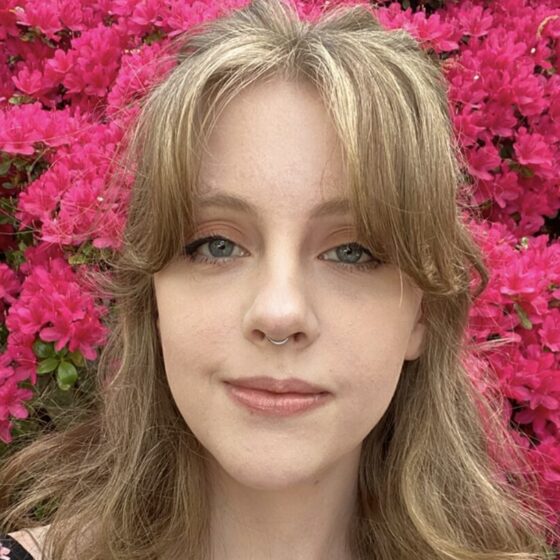 A blond woman with blue eyes and a nose ring. She's standing in front of a pink flowering bush or tree.