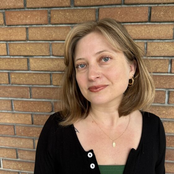 A white woman with blond hair to her shoulders, tilting her head toward the left. She's standing in front of a light-brown-brick wall and wearing a button-down black sweater over a green top, and a necklace
