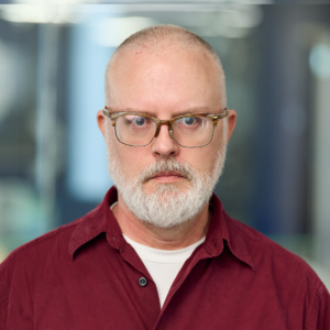 A white man with glasses, a white beard, and very close-cropped hair. He's wearing a button-down maroon shirt over a white T-shirt
