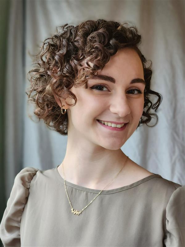 Audrey Baker, a white woman with a brown curly bob, smiles with her head turned to the right. She wears a gold necklace and earrings and a tan blouse with puffed sleeves.