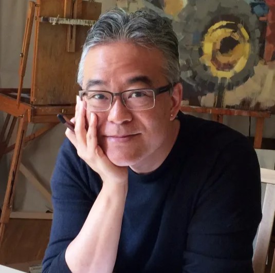 An Asian man with salt-and-pepper hair, resting his chin in his right hand. He's sitting in an art studio and wearing a navy, long-sleeved shirt and rectangular glasses.