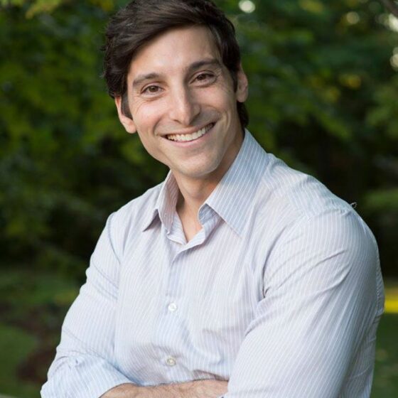 A white man with short, brown hair, folding his arms and smiling. He's standing outside, and wearing a white button-up that is rolled up at the sleeves.