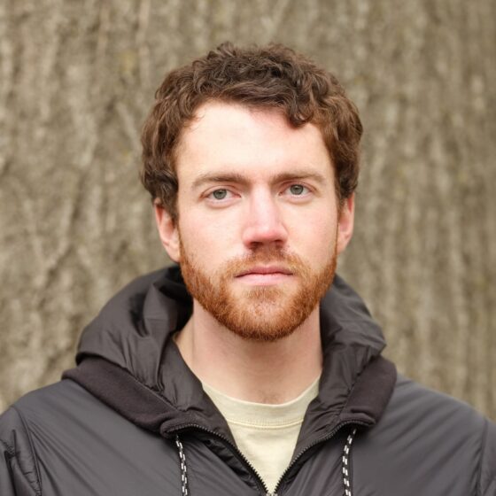 A white man with curly, red hair and a short beard. He is standing in front of a wooden background, and wearing a beige shirt underneath a gray hoodie.