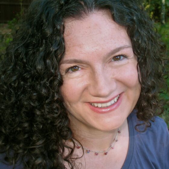 A white woman with long, curly, black hair past her shoulders, sitting on a metal chair and smiling. She's outside and wearing a teal blue shirt and a short, silver necklace adorned with various jewels.