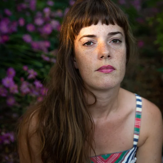 A white woman with long, brown hair and bangs. She's outside, wearing a blue, coral, and white chevron tank top, and looking forward at the camera.