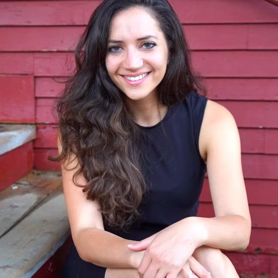 A woman with long, wavy, brown hair, sitting on a bench with her legs crossed and smiling. She is sitting in front the side of a red house, and wearing a black dress.