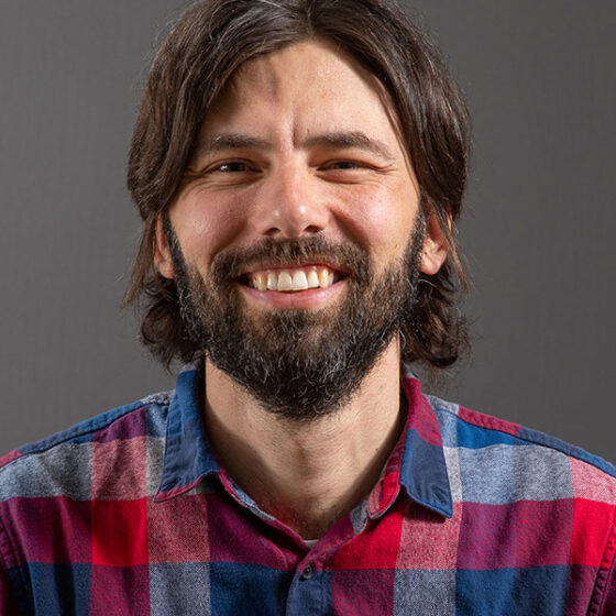 A white man with longer brown hair and a beard, smiling. He is standing in front of a gray background, and wearing a blue-and-red, plaid shirt.