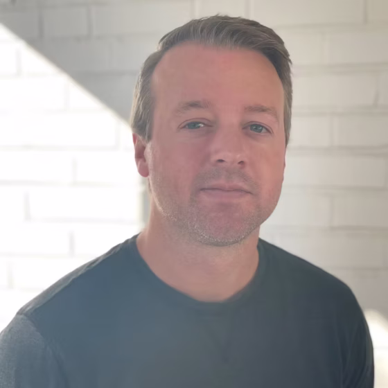 An white man with light-brown hair looking forward at the camera. He's standing in front of a white, brick wall and wearing a black shirt.