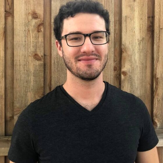 A white man with curly black hair and a stubbly beard, smiling. He stands in front of a wooden fence, and wears a black, v-neck t-shirt and rectangular glasses.