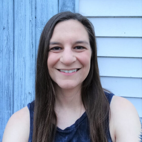 A white woman with brown hair to her chest, looking forward and smiling. She's standing in front of the white wall of a house and wearing a navy tank top.