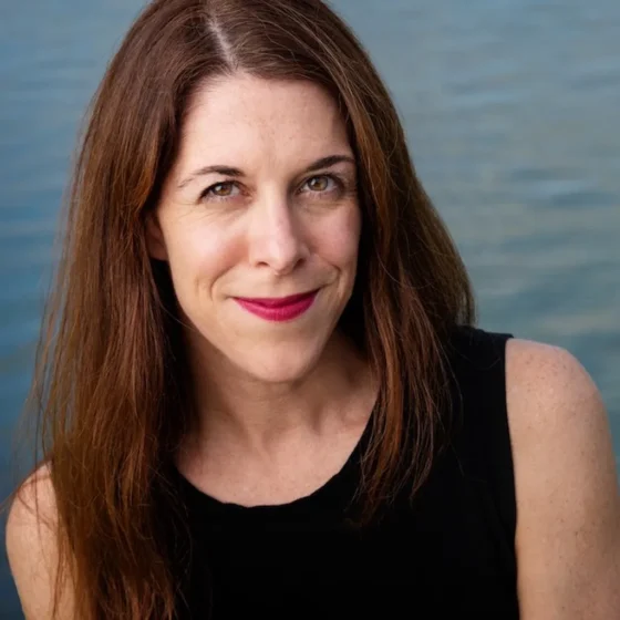 A white woman with straight, brown hair, smiling. She is in front of a blurred, ocean background, and wearing a black tank top and dark, pink lipstick.