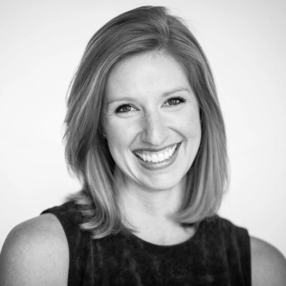 A black-and-white photo of a white woman with hair to her shoulders, smiling at the camera. She's wearing a dark tank top.