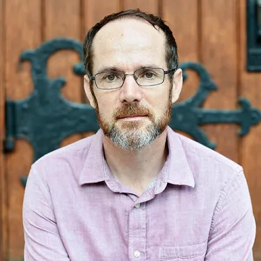 A white man with a gray and brown beard and hair. He is sitting in front of a grand, wooden door, and wearing a pink button-down and rectangular glasses.