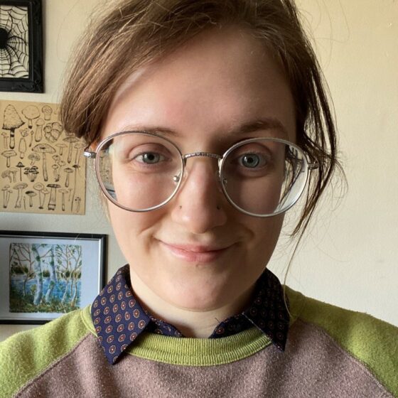 Dana Jean Rider has her hair up and is wearing round, wire-rimmed glasses. She smiles at the camera and is wearing a collared blouse underneath a green and grey sweater.