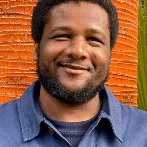 An black man with short black hair, smiling. He's standing in front of an orange wall, and wearing a navy shirt with a blue button up overtop.