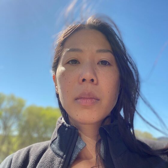 An Asian woman with windswept, black hair past her shoulders, looking down at the camera. She's standing outside and wearing a black running jacket.