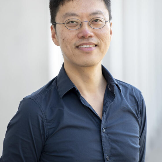 An Asian man with short black hair, smiling. He's standing in front of a white wall, and wearing a navy button-up and oval-shaped glasses.