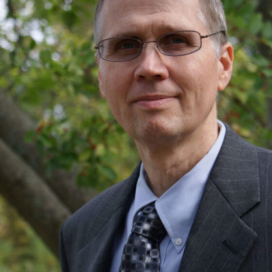 A white man with gray hair, looking beyond the camera and smiling. He is standing outside and wearing a light blue button-up, a navy, patterned tie, a charcoal suit jacket, and rectangular glasses.
