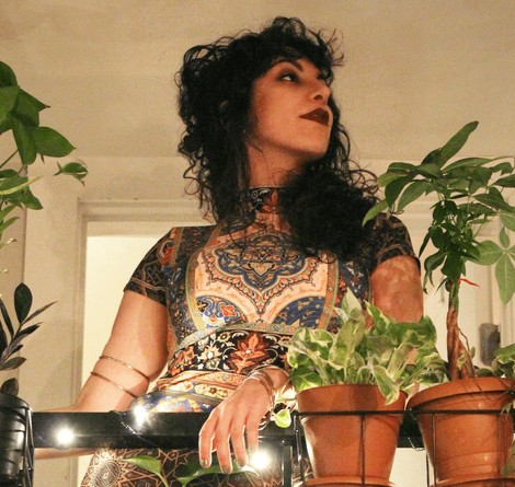 An Iranian-American woman with long, black, curly hair, looking to the right. She is standing in a house with white walls, and has one hand on a black, wire shelf with potted plants on it. She is wearing a tapestry-like dress, golden armbands, and dark pink lipstick.