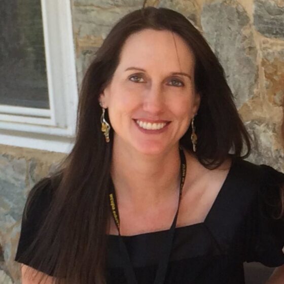 An White woman with brown hair past her shoulders, smiling. She's standing outside of a stone building, wearing a black, short-sleeved blouse and large, gold earrings.