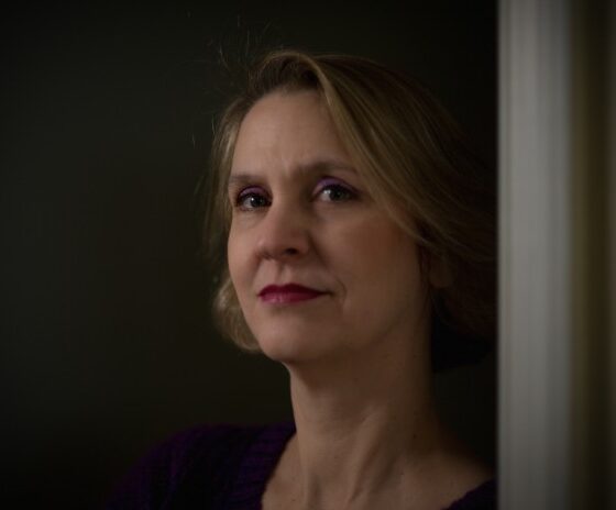 A white woman with blonde hair in a bob, looking at the camera and facing the left. She leans up against a gray wall in front of a black background.