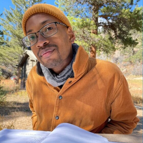 A black person leaning over a table. They are sitting outside at a bench with a notebook, and he is wearing an orange beanie, an orange jacket, a gray scarf, and rectangular glasses.