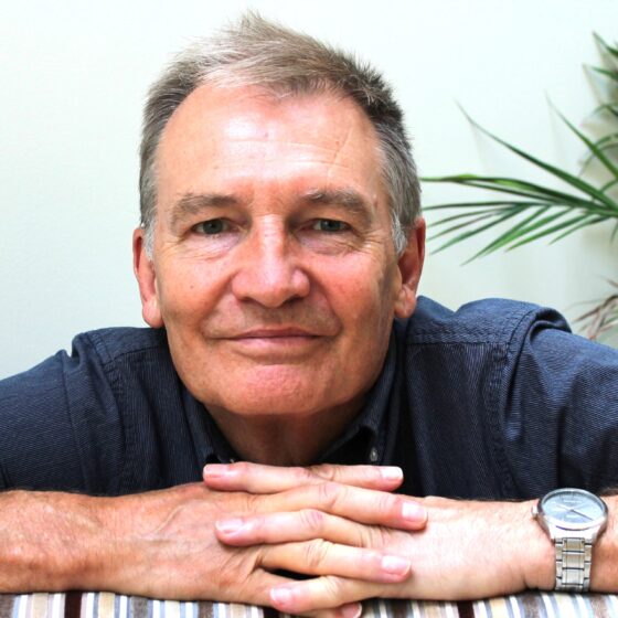An white man with gray hair, leaning on a table and smiling. He's in front of a white wall and the table has a white-and-gray-striped tablecloth on it. He is wearing a navy shirt and a watch on his left wrist.
