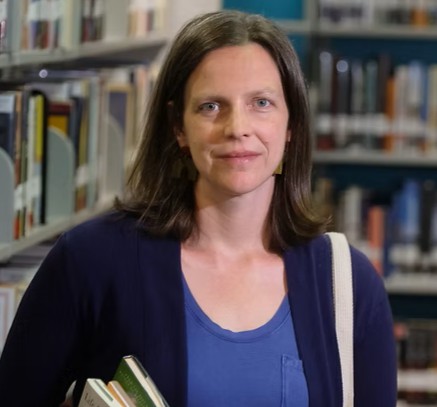 A white woman with brown hair to her shoulders, looking forward. She's standing in a library, holding two books with a white bag over her shoulder, and wearing a blue shirt with a navy cardigan.