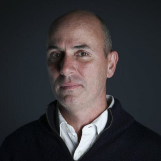 A white man looking forward at the camera. He's standing in front of a black background and wearing a black sweater with a white button-up underneath.