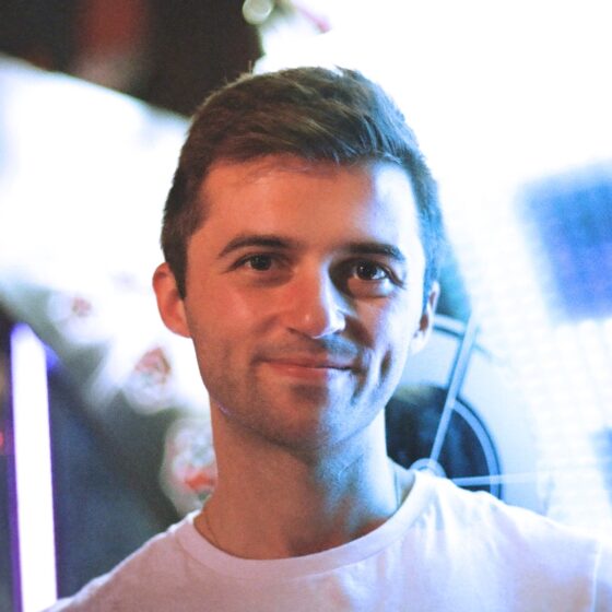 A man with dirty blonde hair and a five-o'clock shadow, smiling. He is standing in front of a blurred city setting with bright lights, and wearing a white t-shirt.