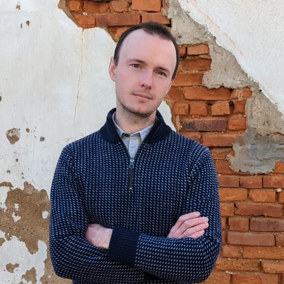 An white man with dark-brown hair with his head tilted to the right and his arms crossed. He's standing in front of a white-plaster and red-brick wall, and wearing a blue button up underneath a navy quarter-zip.