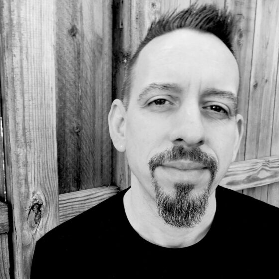 A black-and-white photo of a white man with hair that has been spiked upwards and a goatee. He stands against a wooden background and wears a black shirt.