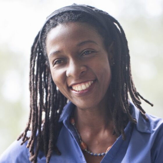 An East African woman with black hair in locs past her shoulders, smiling. She's standing outside, wearing a blue jacket over and a black headband.