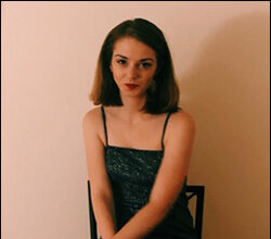 A white woman with brown hair that goes to her shoulders, sitting in a chair and smiling. She's sitting in front of a pink wall, and wearing a black camisole and red lipstick.