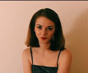 A white woman with brown hair that goes to her shoulders, sitting in a chair and smiling. She's sitting in front of a pink wall, and wearing a black camisole and red lipstick.