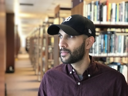 An Asian American man with a short, black beard, looking to the left. He's standing in a library, and wearing a dark, maroon button up and a black Detroit Tigers hat.