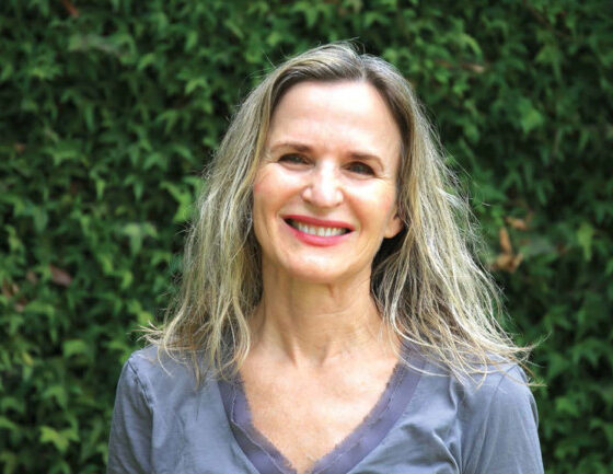 A white woman with wavy, blonde hair, smiling. She is standing in front of a bush background, wearing a gray v-neck t-shirt.