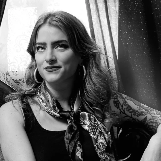 A black-and-white photo of a white woman with long, wavy hair. Her left elbow on the back of the couch and her right hand is holding her right knee as she crosses her legs. She is sitting on an old-fashioned couch with curtains in the background. She is wearing a black tank top, black jeans, a patterned scarf tied around her neck, and hoop earrings.