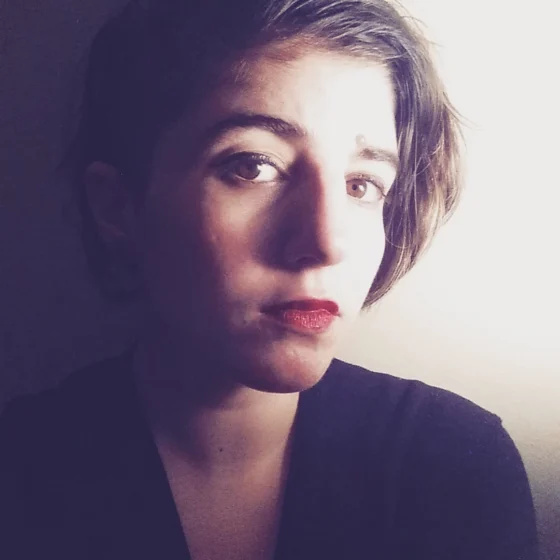 A white woman with brown hair just past her chin, tilting her head to the right. She's standing against a white wall, wearing a black, long-sleeved shirt.