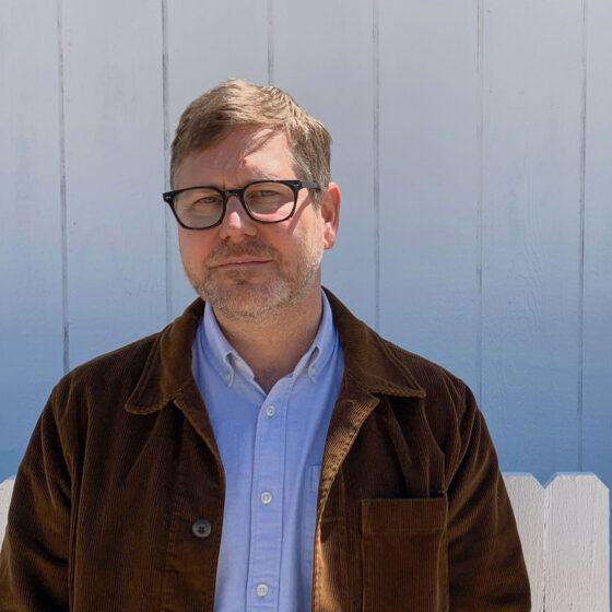 An white man with light-brown hair and a short, brown beard looking forward at the camera. He's standing in front of a white fence and wearing a light blue button down with a brown corduroy jacket overtop, with black, square glasses.