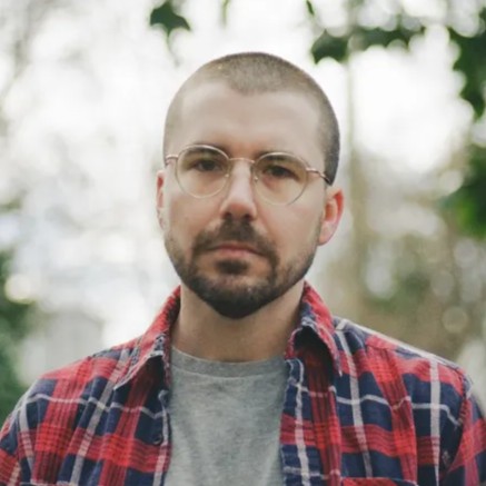 An white man with a short, black beard looking forward at the camera. He's standing outside and wearing a gray shirt with a red and navy flannel overtop, with round glasses.