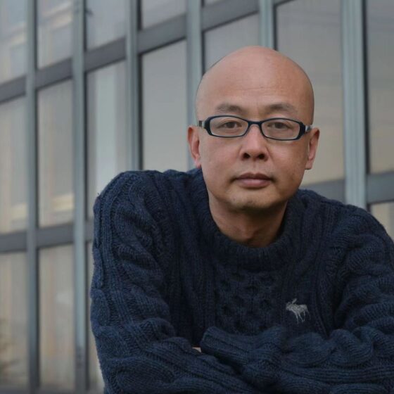 A bald, Asian man. He's standing in front of the windows of a glass building, and wearing a navy sweater and rectangular glasses.