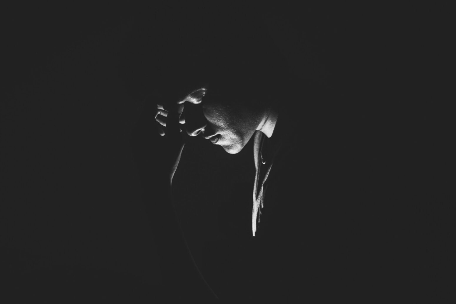 A dark room with one light shining on a man's face. The man looks down with a solemn expression, pressing his thumb and index finger into the bridge of his nose with his eyes closed.
