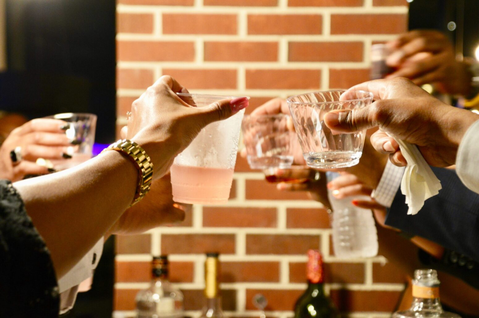 Several hands holding up different kinds of glasses with drinks in them, in a toast. The background is a brick wall column, with a few liquor/wine bottles next to it
