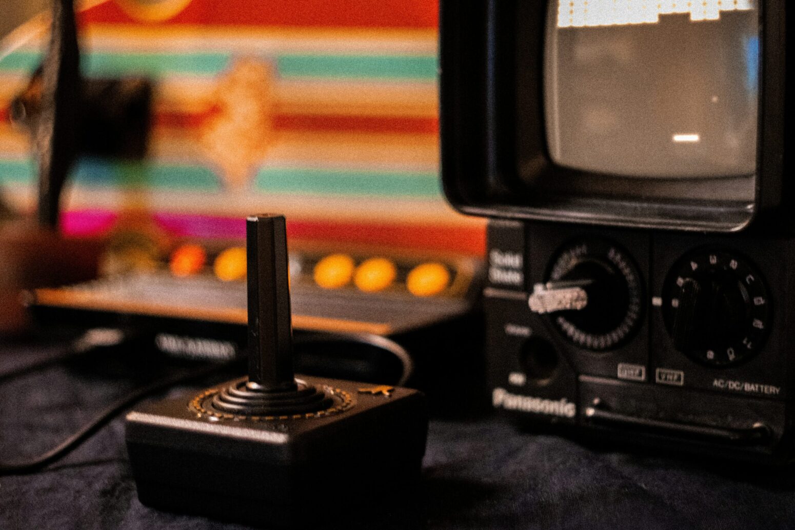An Atari controller, hooked up to a small, old-fashioned monitor, sitting on a black table. In the background, a red, blue, and white-striped display.