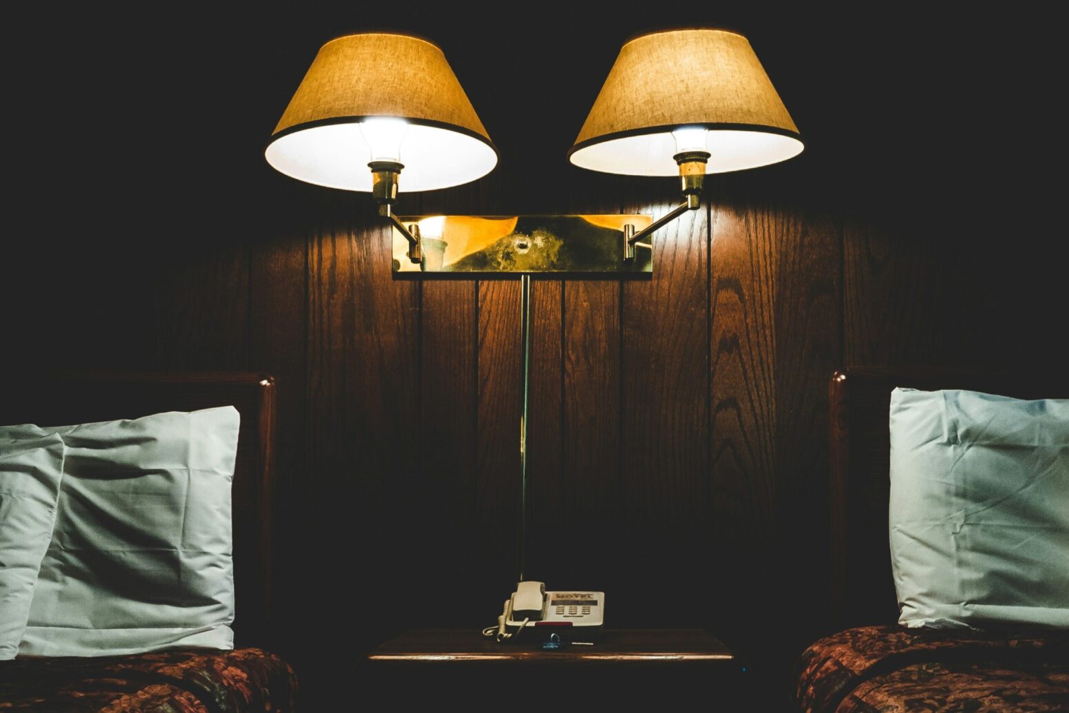An old motel room with wooden-panel walls. Two sconces alight a beside table with
