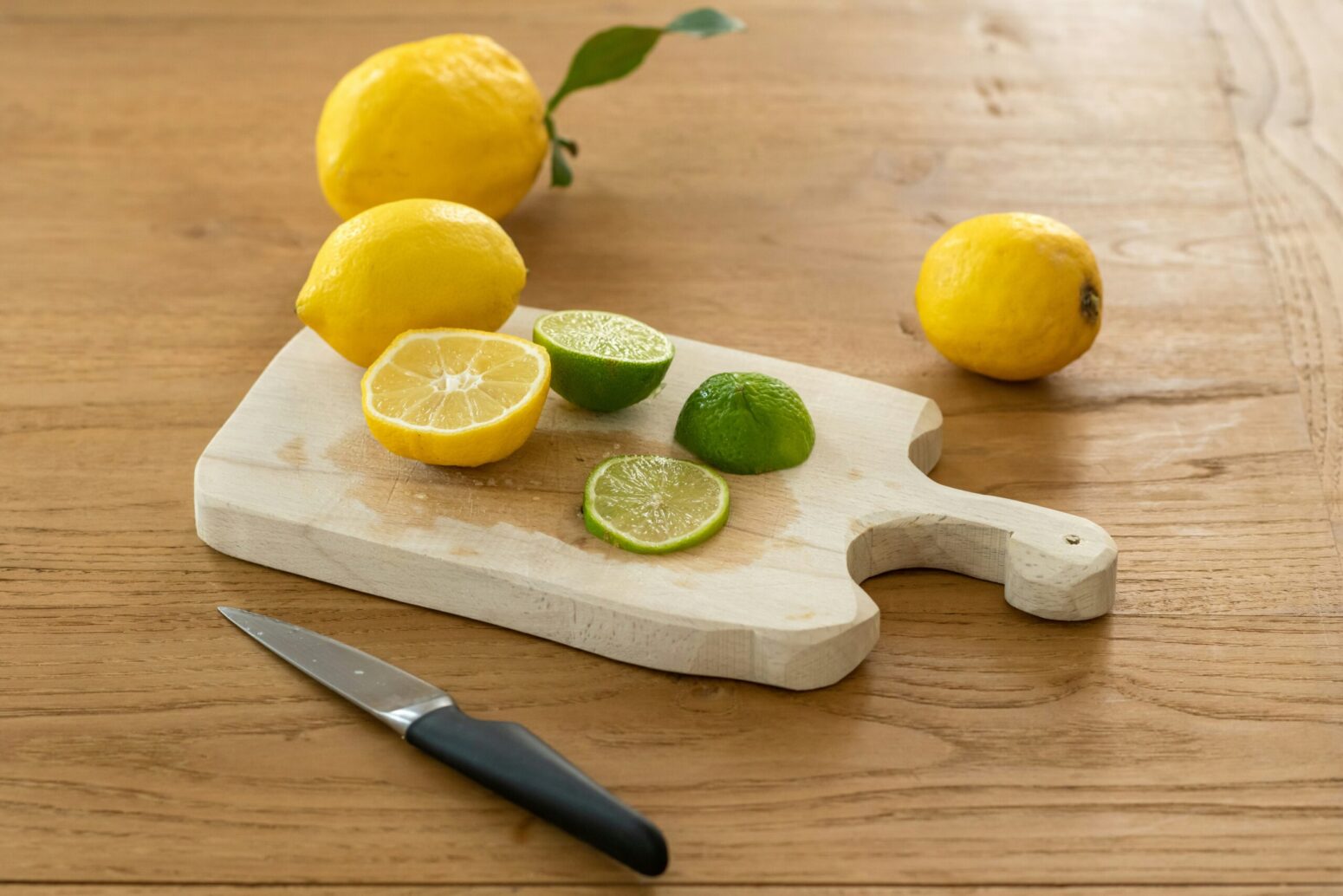 A wooden cutting board on a table. On the board are some lemons and limes. A small cutting knife lays next to it.