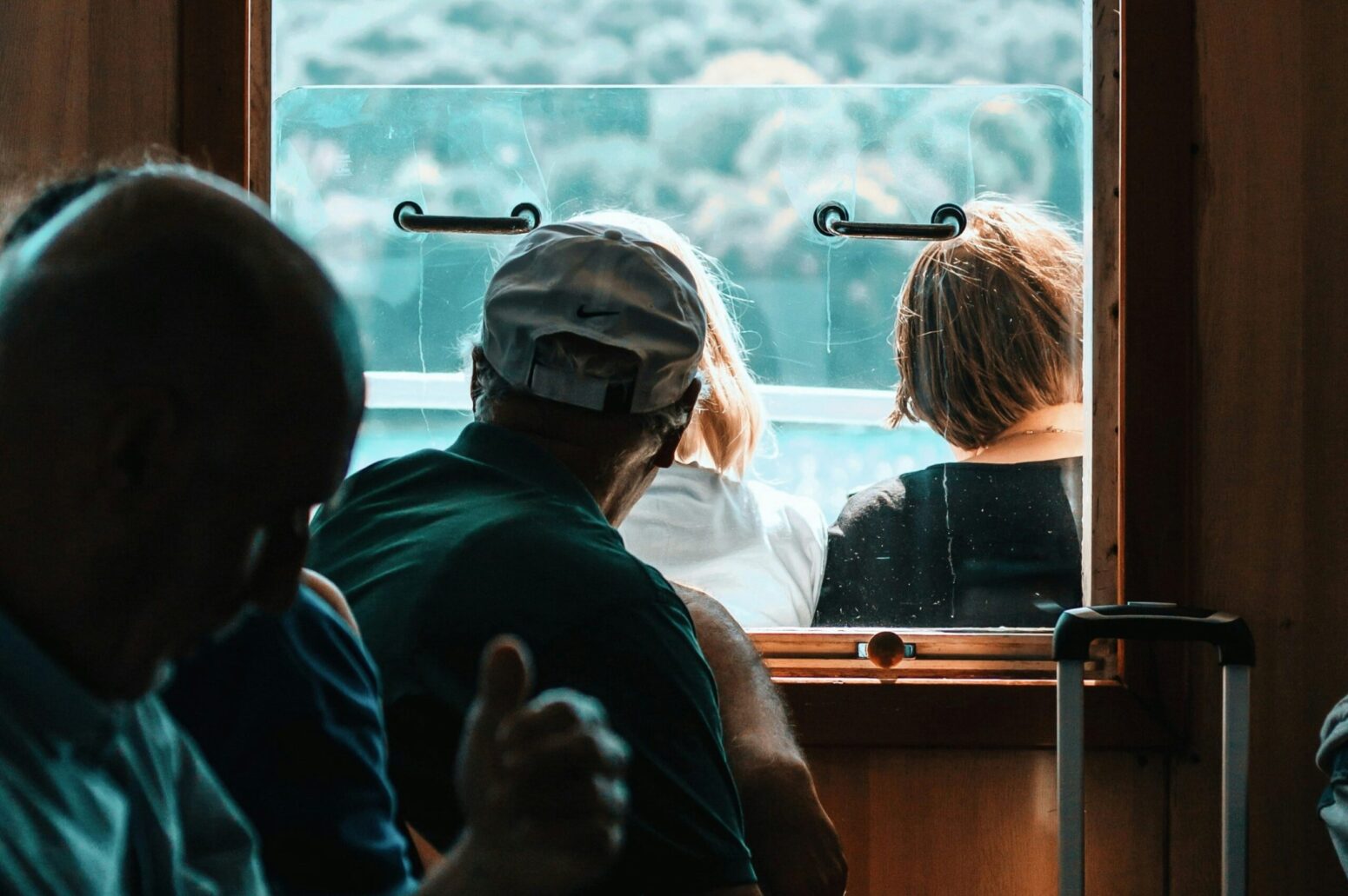 A man in a hat looks out the window of a steamboat. Another man sits to his right, looking down. Two women sit outside together. The view from the steamboat is of a large forest.