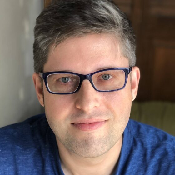 A white man with black and gray hair, and blue eyes. He's wearing glasses and wearing a royal blue polo.